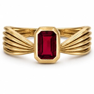 May include: A gold ring with a rectangular, deep red gemstone at the center. The ring's band features a layered design that flows outward from the gemstone. The ring is set against a white background.