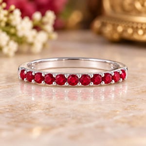 May include: A silver-toned band ring featuring a row of eleven round-cut, vibrant red gemstones set in a shared-prong setting. The ring has a polished metallic finish and a slim, uniform profile that spans the width of the band.