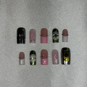 Könnte beinhalten: Eine Sammlung von zehn künstlichen Nägeln in verschiedenen Designs. Die Nägel weisen Farben wie Schwarz, Rosa, Weiß und Grün auf, wobei einige mit silbernen Akzenten und Text verziert sind. Die Designs umfassen Muster, Text und ein Metallic-Finish.