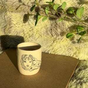 Puede incluir: Una taza de cerámica de color crema con un dibujo lineal negro de una persona con el pelo largo y fluido adornado con pequeñas estrellas. La taza tiene una textura moteada y una forma cilíndrica como recipiente para bebidas.