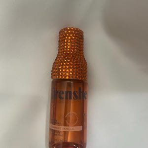 Puede incluir: Una pequeña botella transparente de bruma corporal con una tapa naranja enjoyada. La botella contiene un líquido marrón y una etiqueta que dice "Cashmere Vanilla". La marca "frenshe" está impresa en la botella.