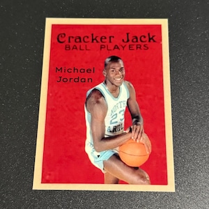 May include: A vintage Cracker Jack trading card featuring Michael Jordan in a North Carolina Tar Heels basketball uniform. The card has a red background with the text "Cracker Jack Ball Players" and "Michael Jordan".