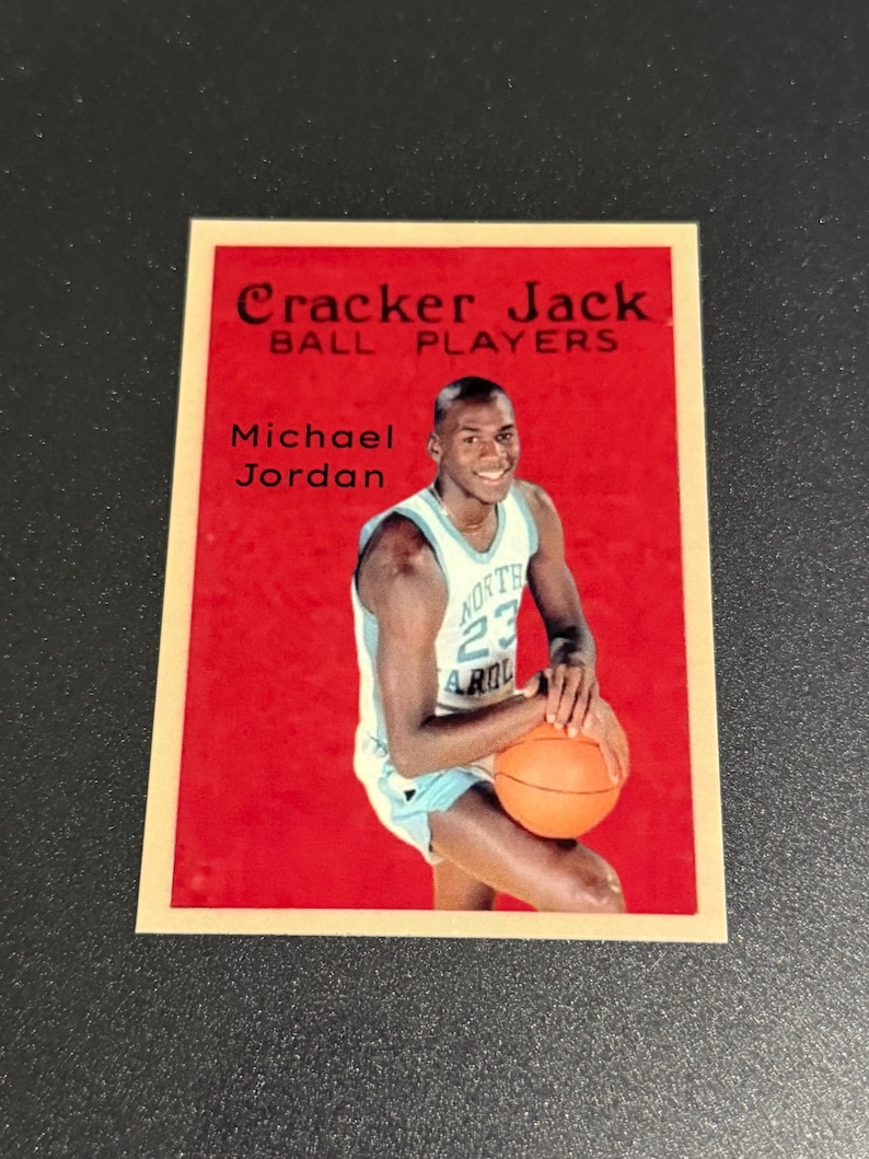 May include: A vintage Cracker Jack trading card featuring Michael Jordan in a North Carolina Tar Heels basketball uniform. The card has a red background with the text "Cracker Jack Ball Players" and "Michael Jordan".
