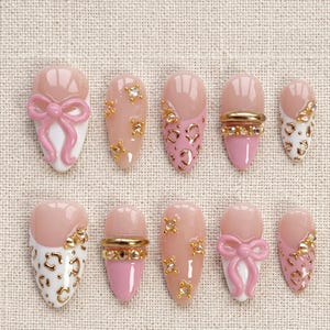 May include: A set of ten artificial nails in various designs. The nails feature a combination of pink, white, and gold accents. Some nails have bow details, while others have gold rhinestones or leopard print patterns. The nails are oval-shaped.