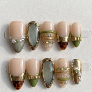 May include: A set of ten press-on nails featuring a nude base with olive green tips, gold metallic accents, and small embedded pearls and crystals. The designs include tortoiseshell patterns, textured gold bands, and smooth, glossy finishes.
