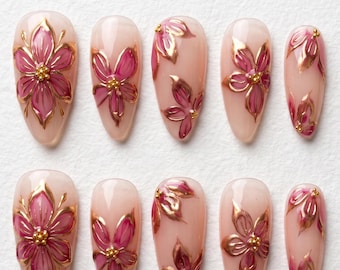 May include: A set of ten almond-shaped press-on nails with a translucent nude base. Each nail features hand-painted pink floral designs outlined in metallic gold paint, accented with small gold beads at the center of each flower.