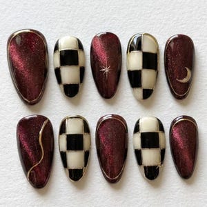 May include: A set of ten press-on nails featuring a mix of deep burgundy cat-eye magnetic polish and black and white checkered patterns. Some nails include gold metallic accents, such as a thin curved line, a small star, and a crescent moon.