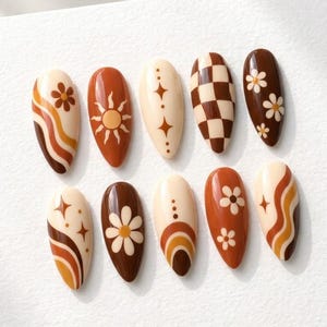 May include: A set of ten almond-shaped press-on nails featuring a color palette of cream, tan, orange, and dark brown. Designs include wavy lines, a sun motif, four-pointed stars, a brown and cream checkerboard pattern, and white daisy flowers.