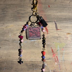 May include: A keychain featuring a gold-tone lobster clasp, a central square acrylic charm with a dark red forest image, and two dangling beaded strands. The strands include pink, purple, and red beads, a small black tassel, and star charms.