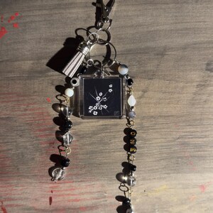 May include: A keychain featuring a silver lobster clasp, a light gray tassel, and a square charm with a black and white floral design and the text ΦΜΣ. Two beaded strands hang from the charm, incorporating clear, black, and white beads, gold letter beads spelling ONE, and a small dragonfly charm at the end.