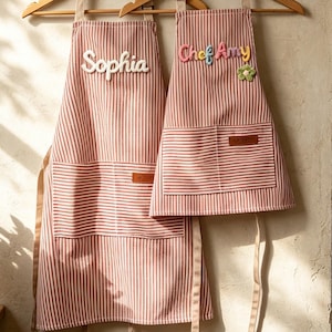 Könnte beinhalten: Zwei passende Schürzen mit rot-weißem vertikalem Streifenmuster, die an Holzkleiderbügeln hängen. Die größere Schürze trägt den weißen Schriftzug "Sophia" und die kleinere den bunten Schriftzug "Chef Amy" mit einer kleinen grünen Blumenapplikation.