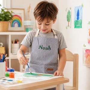 Könnte beinhalten: Eine marineblau-weiß gestreifte Schürze mit dem Namen Noah, der in grünem Garn auf die Brust gestickt ist. Die Schürze verfügt über weiße Nackenbänder und eine Vordertasche mit einem kleinen braunen rechteckigen Aufnäher am unteren Rand.