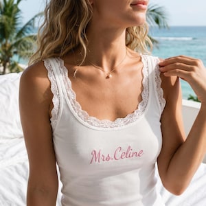 Könnte beinhalten: Ein weißes, geripptes Tanktop mit zarter weißer Spitzenborte am Rundhalsausschnitt und an den Trägern. Auf der Vorderseite des Kleidungsstücks ist der Text Mrs. Celine in einer zartrosa Schreibschrift mittig auf der Brust eingestickt.