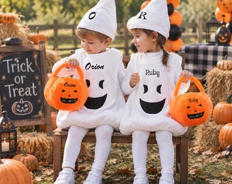 May include: Two children wearing white ghost costumes with black facial features and matching pointed hats. One hat features the letter O and the other the letter R. Each child holds an orange plush pumpkin bucket labeled with the names Orion and Ruby.