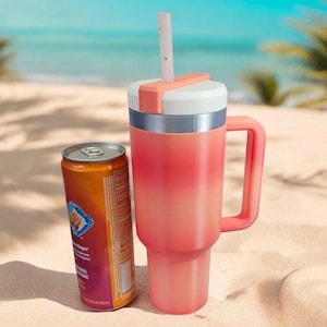 May include: A coral-colored insulated tumbler with a handle and a white lid, featuring a matching straw. Beside it is an orange can with a blue and white label. The items are on a sandy beach with the ocean in the background.