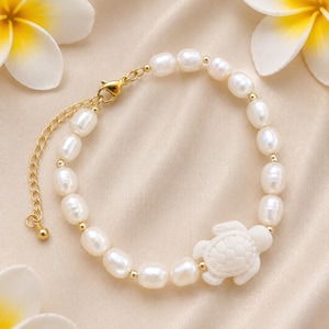 May include: A delicate bracelet featuring white oval pearls and small gold beads. A white turtle charm is centered on the bracelet. The bracelet has a gold chain and clasp. The background is a soft, neutral color.