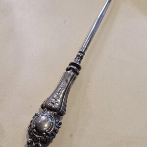Puede incluir: Un gancho para botones de metal plateado con un mango ornamentado y en relieve con intrincados detalles de volutas y motivos florales. El eje largo y delgado termina en un pequeño gancho curvo diseñado para abrochar botones en la ropa.