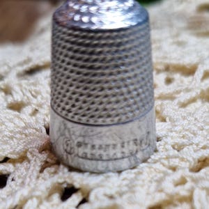 May include: A silver-colored metal thimble featuring a textured, cross-hatched pattern on the upper dome and sides. The base of the thimble has a smooth, polished band with engraved text that appears to read 'Chester' and 'England'.