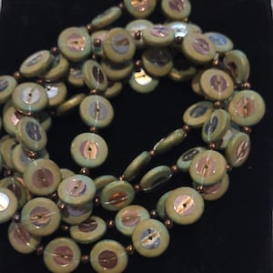May include: A necklace composed of multiple circular, sage green ceramic beads, each featuring a central, iridescent, shell-like inlay. The beads are strung together with small, metallic copper-colored spacer beads along a thin, clear filament.