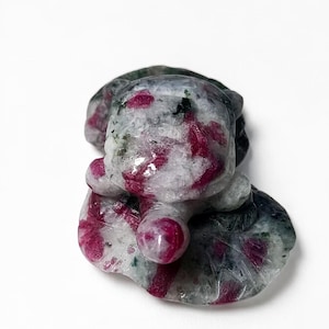 Pode incluir: Uma estatueta de pedra esculpida de uma tartaruga sobre uma folha de vitória-régia, feita de um mineral polido com uma base branca e cinzenta manchada, com inclusões proeminentes de um vermelho magenta vibrante na superfície.