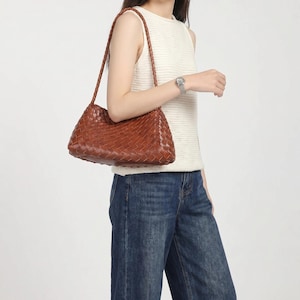 Può includere: Una borsa a spalla in pelle marrone caratterizzata da una trama intrecciata e un manico intrecciato. La borsa ha una forma rettangolare con angoli arrotondati e una finitura liscia, con un motivo intrecciato uniforme su tutta la superficie.