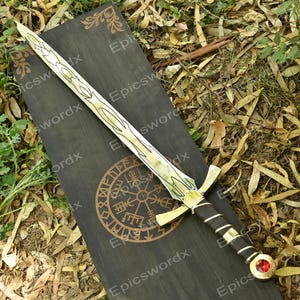 Peut inclure: Une épée décorative avec une lame et une garde dorées, avec une poignée noire et marron. La lame présente un motif blanc et vert. L'épée repose sur une surface en bois noir avec un emblème doré et le texte "Epicswordx".