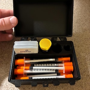 May include: A black plastic storage case with red latches containing several insulin syringes with orange caps, a stack of Embecta alcohol prep pads, and a yellow circular container. The syringes feature clear barrels with black measurement markings.