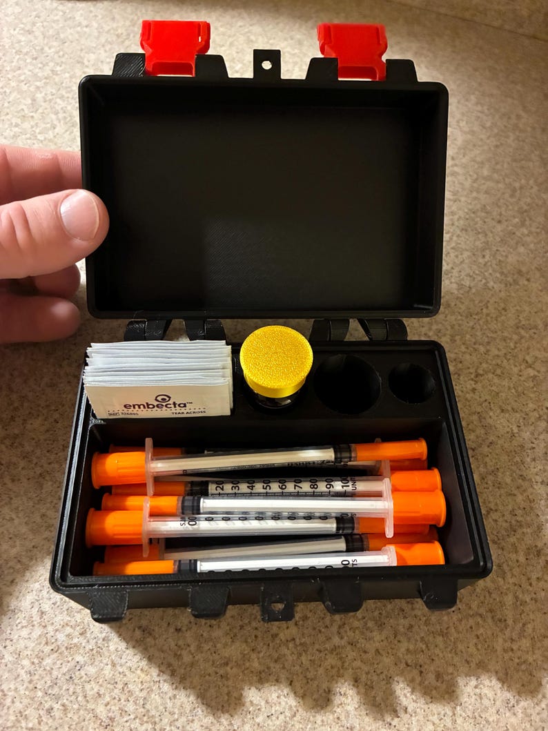 May include: A black plastic storage case with red latches containing several insulin syringes with orange caps, a stack of Embecta alcohol prep pads, and a yellow circular container. The syringes feature clear barrels with black measurement markings.
