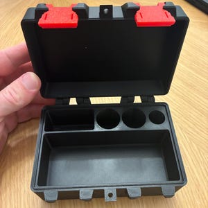 May include: A black rectangular storage case with a hinged lid and two bright red latches. The interior features a large bottom compartment, a smaller rectangular section, and three circular cylindrical slots for organised storage.