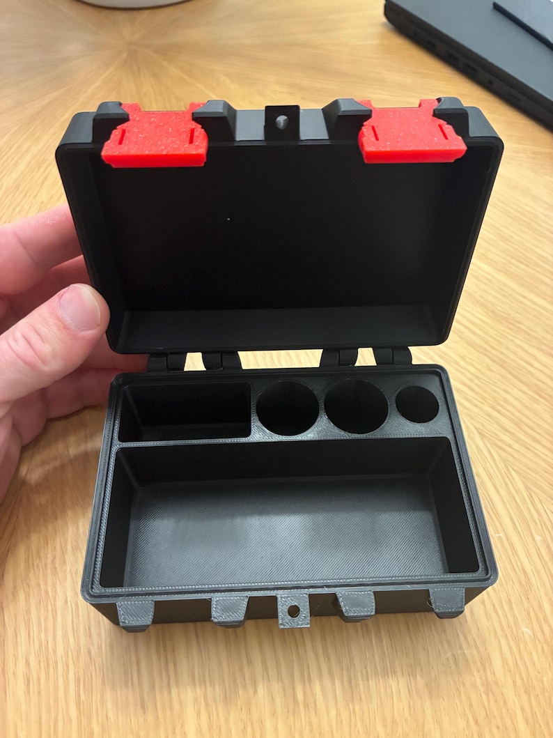 May include: A black rectangular storage case with a hinged lid and two bright red latches. The interior features a large bottom compartment, a smaller rectangular section, and three circular cylindrical slots for organised storage.