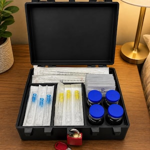 Puede incluir: Un estuche de almacenamiento r&iacute;gido, negro y rectangular, con un candado rojo y una llave. El estuche abierto contiene suministros m&eacute;dicos, incluyendo cuatro viales de vidrio con tapas azules, agujas envueltas individualmente y jeringas.