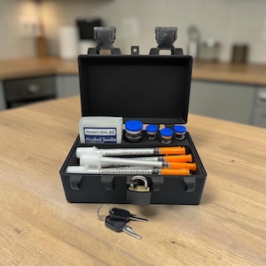 May include: A black plastic storage case with two latches and a small padlock, containing four syringes with orange caps, four small vials with blue caps, and a box of Member's Mark alcohol swabs. Two metal keys are placed in front of the case.