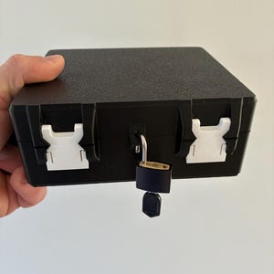 Puede incluir: Una caja de almacenamiento rectangular negra con superficie texturizada, que cuenta con dos hebillas laterales de pl&aacute;stico blanco y un peque&ntilde;o candado de lat&oacute;n con una llave insertada en el cierre central para asegurar la tapa.