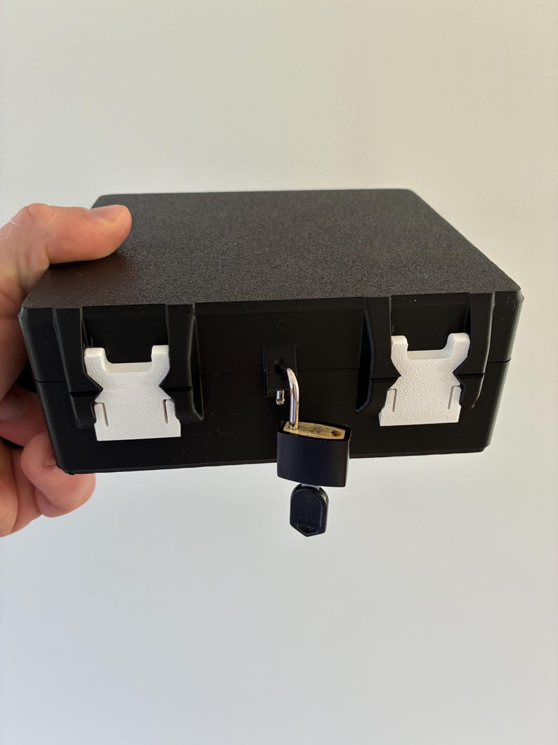 Puede incluir: Una caja de almacenamiento rectangular negra con superficie texturizada, que cuenta con dos hebillas laterales de pl&aacute;stico blanco y un peque&ntilde;o candado de lat&oacute;n con una llave insertada en el cierre central para asegurar la tapa.