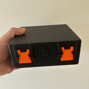 Puede incluir: Un estuche de almacenamiento rectangular negro con un acabado de superficie texturizado. El estuche cuenta con dos cierres de pl&aacute;stico de color naranja brillante en la parte frontal para un cierre seguro y un peque&ntilde;o orificio central.
