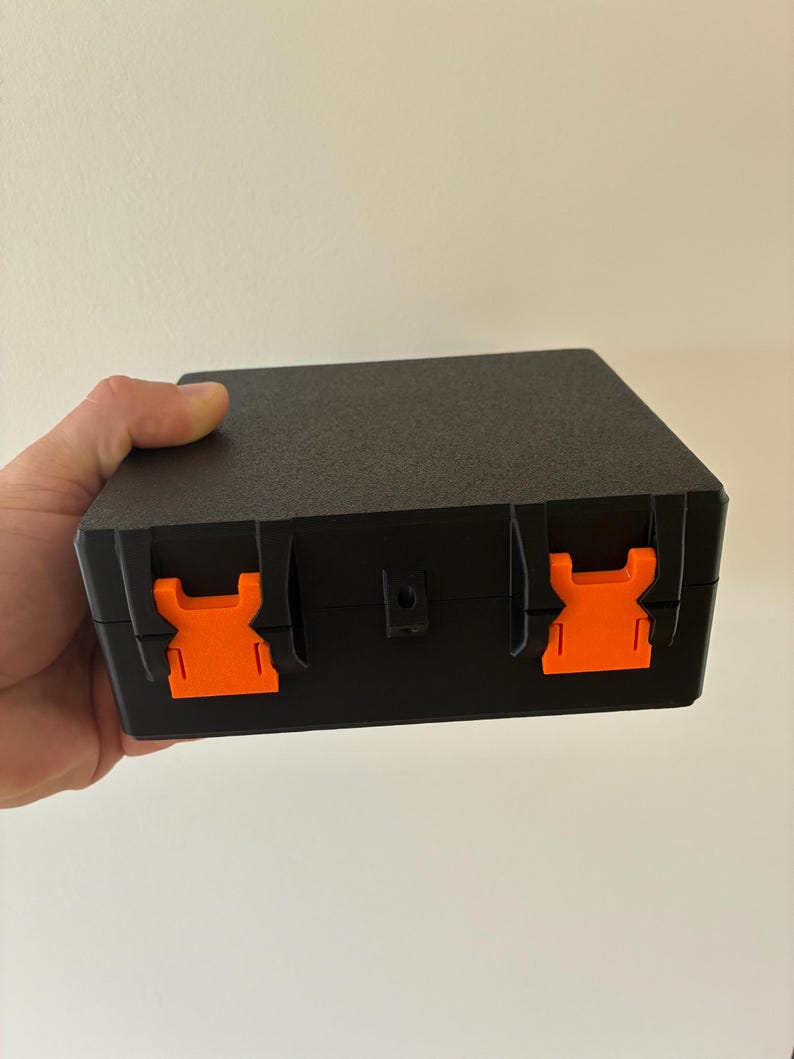 Puede incluir: Un estuche de almacenamiento rectangular negro con un acabado de superficie texturizado. El estuche cuenta con dos cierres de pl&aacute;stico de color naranja brillante en la parte frontal para un cierre seguro y un peque&ntilde;o orificio central.