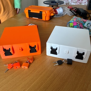 Puede incluir: Dos cajas de almacenamiento cuadradas, una de color naranja brillante y otra blanca, cada una con dos cierres de pl&aacute;stico negro en la parte frontal. Cada caja viene acompa&ntilde;ada de un peque&ntilde;o candado y un juego de llaves para su cierre.