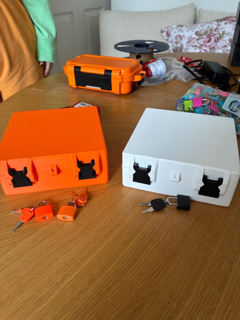 Puede incluir: Dos cajas de almacenamiento cuadradas, una de color naranja brillante y otra blanca, cada una con dos cierres de pl&aacute;stico negro en la parte frontal. Cada caja viene acompa&ntilde;ada de un peque&ntilde;o candado y un juego de llaves para su cierre.