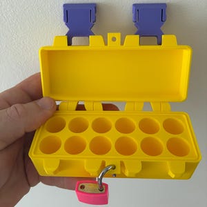 May include: A bright yellow rectangular storage case with a hinged lid and two purple plastic buckles at the top. The interior features twelve circular compartments arranged in two rows. A small pink padlock is attached to the front latch mechanism.