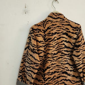 May include: A long-sleeved jacket featuring a tan and black tiger stripe pattern. The garment has a high collar and cuffs with a black and white vertical pinstripe design. The jacket is displayed hanging on a wire hanger against a plain wall.