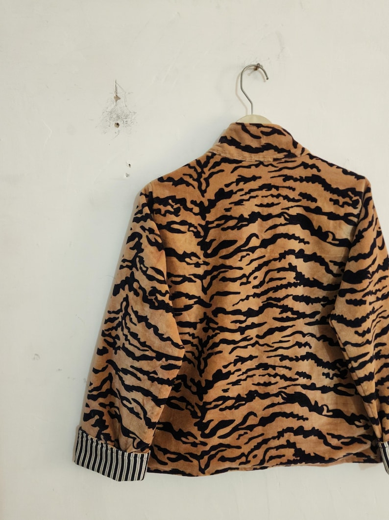 May include: A long-sleeved jacket featuring a tan and black tiger stripe pattern. The garment has a high collar and cuffs with a black and white vertical pinstripe design. The jacket is displayed hanging on a wire hanger against a plain wall.