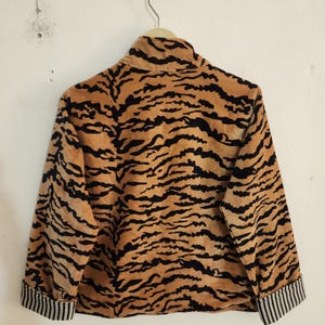 May include: A long-sleeved jacket featuring a tan and black tiger stripe pattern. The garment has a high collar and cuffs with a black and white vertical pinstripe design. The jacket is displayed on a hanger against a plain white wall.