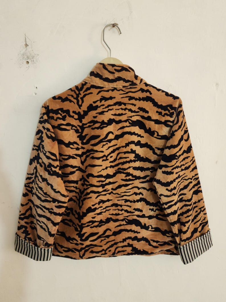 May include: A long-sleeved jacket featuring a tan and black tiger stripe pattern. The garment has a high collar and cuffs with a black and white vertical pinstripe design. The jacket is displayed on a hanger against a plain white wall.