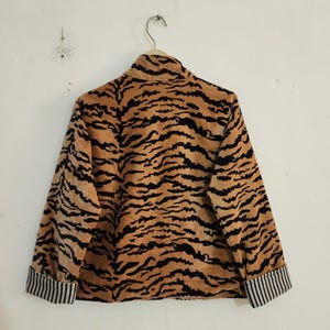 May include: A long-sleeved jacket featuring a tan and black tiger stripe pattern throughout. The garment has a high collar and cuffs that display a contrasting black and white vertical pinstripe design on the interior fold.