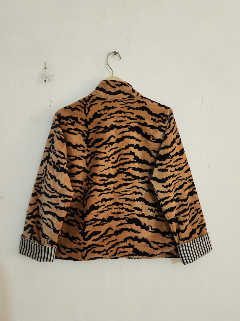 May include: A long-sleeved jacket featuring a tan and black tiger stripe pattern throughout. The garment has a high collar and cuffs that display a contrasting black and white vertical pinstripe design on the interior fold.
