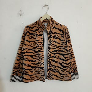 May include: A long-sleeved jacket featuring a tan and black tiger stripe pattern on the exterior. The garment has a high collar, a front opening, and cuffs that reveal a black and white vertical pinstripe lining on the interior fabric.