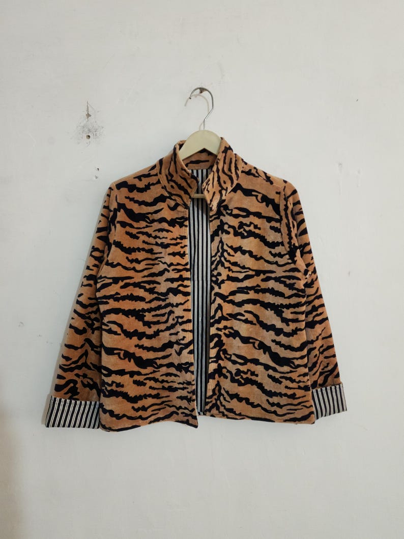 May include: A long-sleeved jacket featuring a tan and black tiger stripe pattern on the exterior. The garment has a high collar, a front opening, and cuffs that reveal a black and white vertical pinstripe lining on the interior fabric.