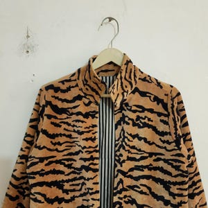 May include: A long-sleeved jacket featuring a tan and black tiger stripe pattern. The jacket has a high collar and is open at the front, revealing a vertical black and white pinstriped lining on the interior. The garment is displayed on a hanger.