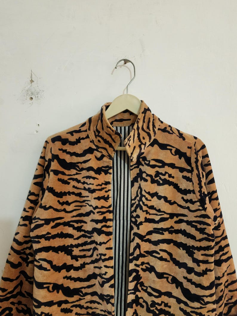 May include: A long-sleeved jacket featuring a tan and black tiger stripe pattern. The jacket has a high collar and is open at the front, revealing a vertical black and white pinstriped lining on the interior. The garment is displayed on a hanger.