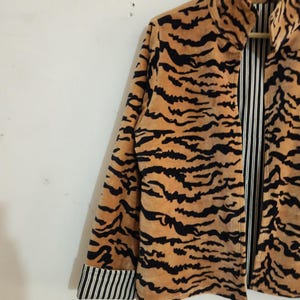 May include: A long-sleeved jacket featuring a tan and black tiger stripe pattern on the exterior. The interior lining and the folded cuffs display a contrasting black and white vertical pinstripe design. The jacket has a high collar and a front opening.
